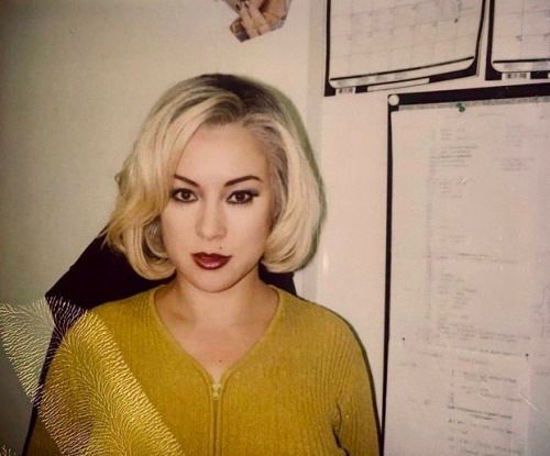 a woman with blonde hair and green eyes posing in front of a wall mounted calendar