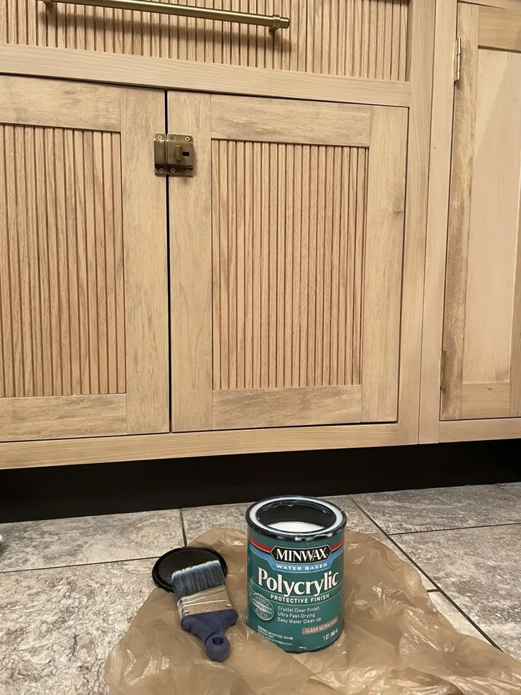 a can of paint sitting on the floor next to a brush and some brown cabinets