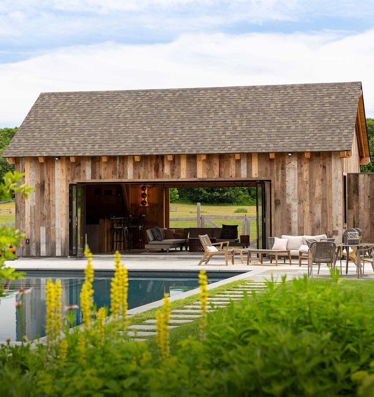 a barn with a pool and patio furniture in the foreground, surrounded by greenery