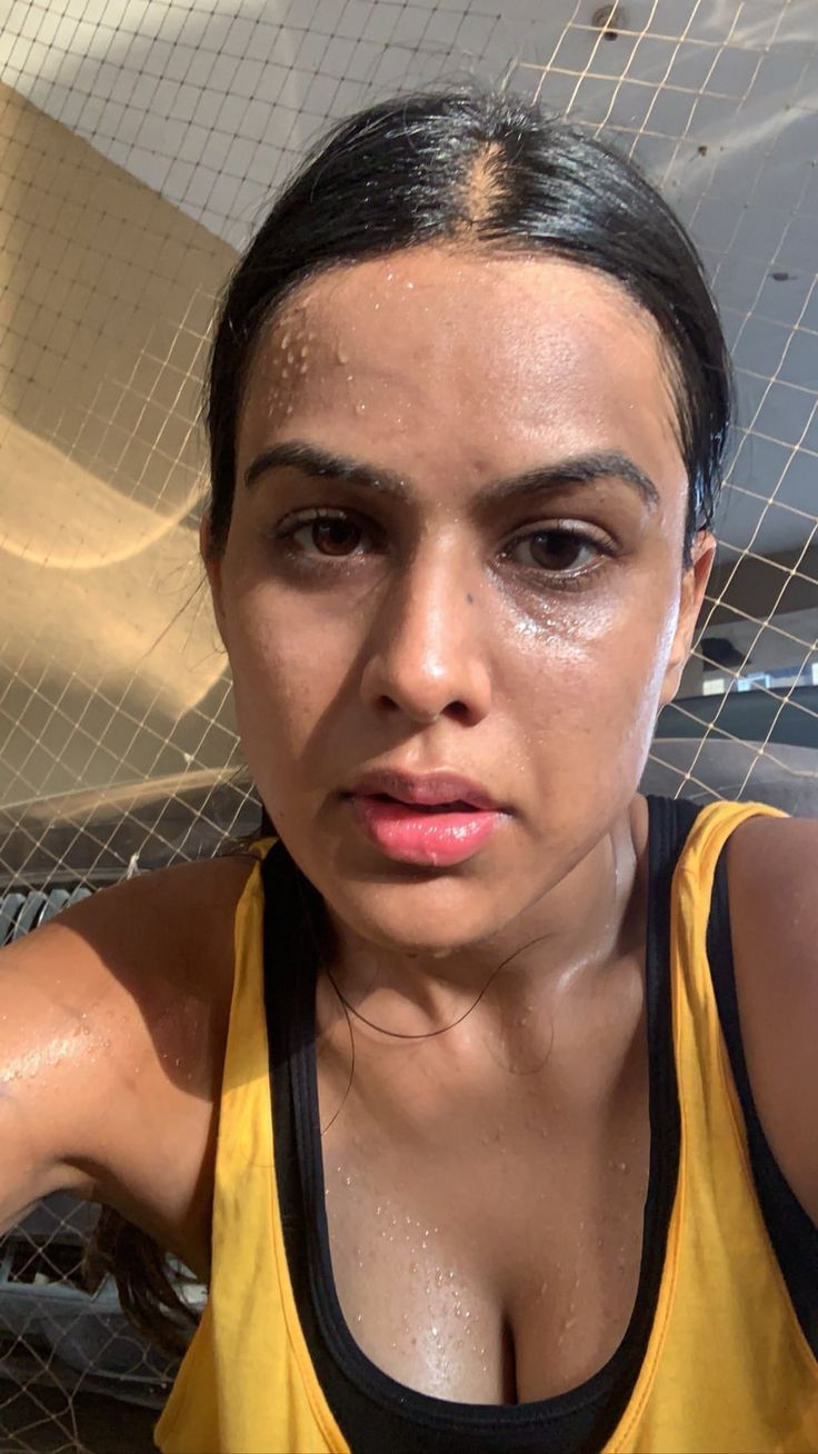 a woman with wet face and yellow tank top looking at the camera while standing in front of a fence