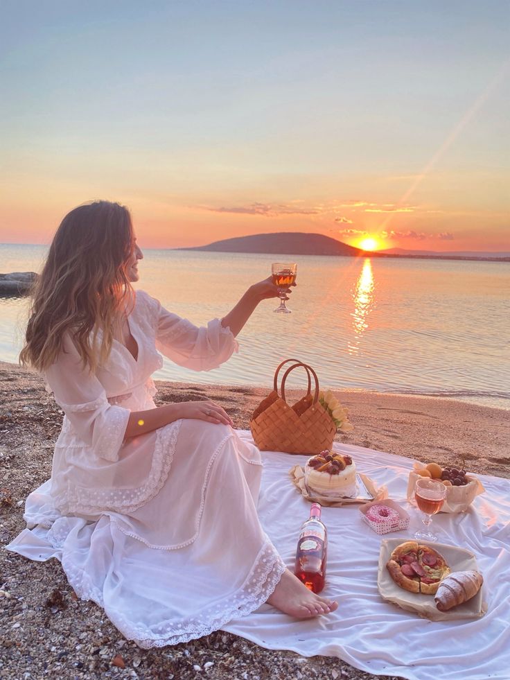 a woman sitting on top of a beach next to the ocean holding a glass of wine