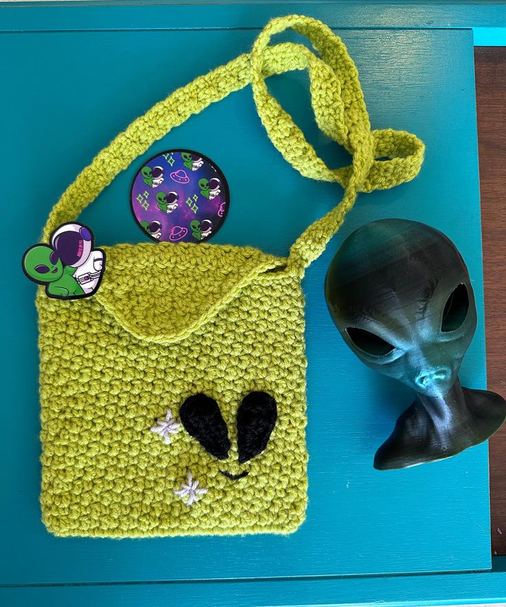 a crocheted bag with an alien head on the side and a plastic skull sitting next to it