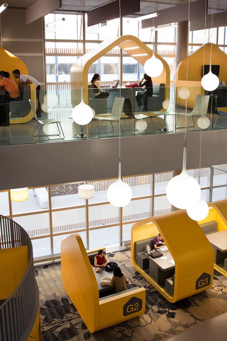 people are sitting at desks in an office with yellow and gray furniture, large windows, and hanging lights