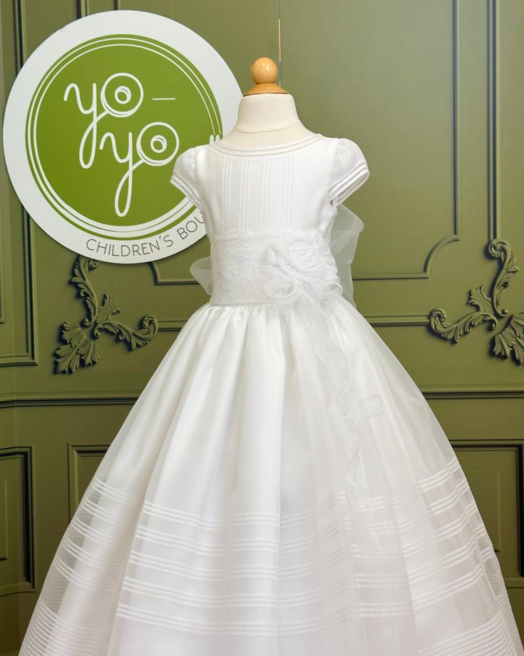 A traditional and classical first communion gown for girls, made with lightweight white organza. It has beautiful pleats throughout the body and skirt. The flower appliqué is removable. It has buttons on the back for closure. Made in Spain Dry clean Does not include crinoline 100% polyester Final Sale Fitted First Communion Dress, Classic Fitted First Communion Dress, First Communion Dress With Ruffles And Fitted Bodice, Classic White First Communion Gown, Classic White Gown For First Communion, Holy Communion Dresses, First Communion Dress, First Communion Dresses, Gowns For Girls