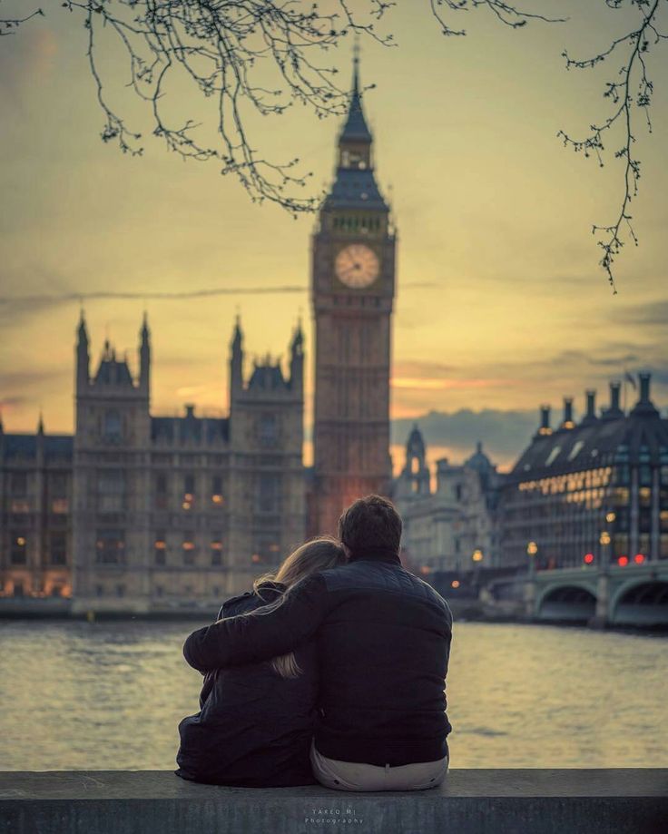 two people sitting on a bench looking at the water and clock tower in the background