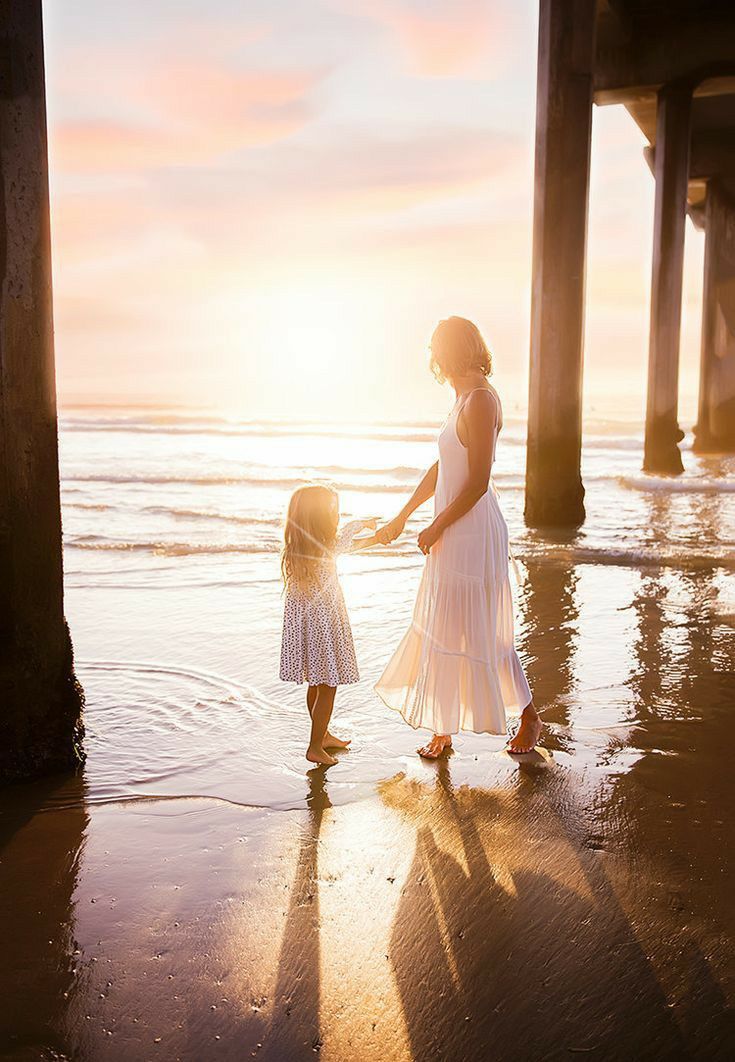 a mother and daughter hold hands as they walk along the beach under a pier at sunset