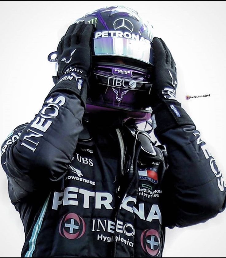 a man in a black jacket and helmet covering his face with both hands while standing next to a white wall