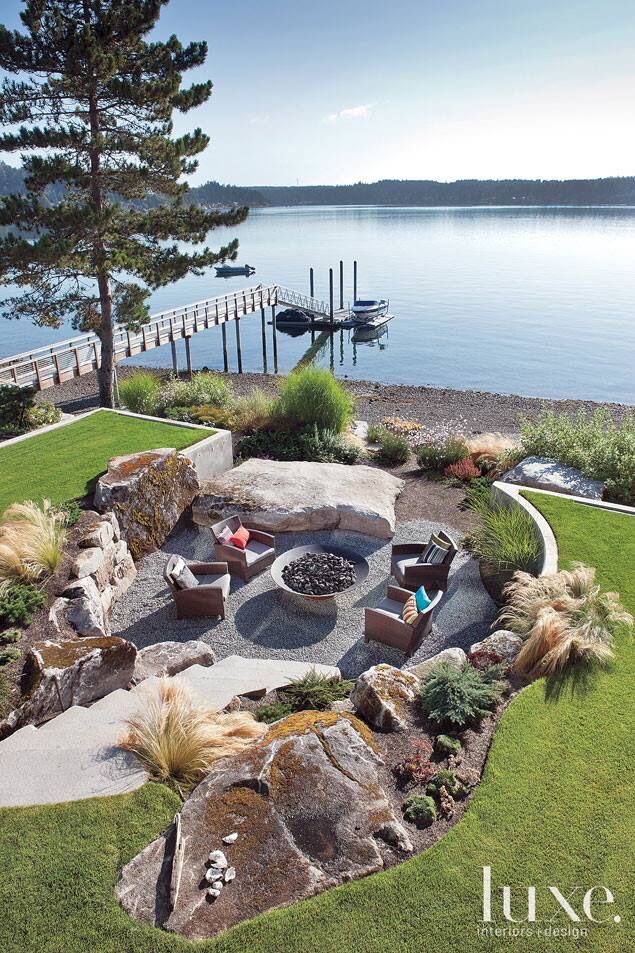 an outdoor fire pit surrounded by rocks and grass next to the water with a dock in the background