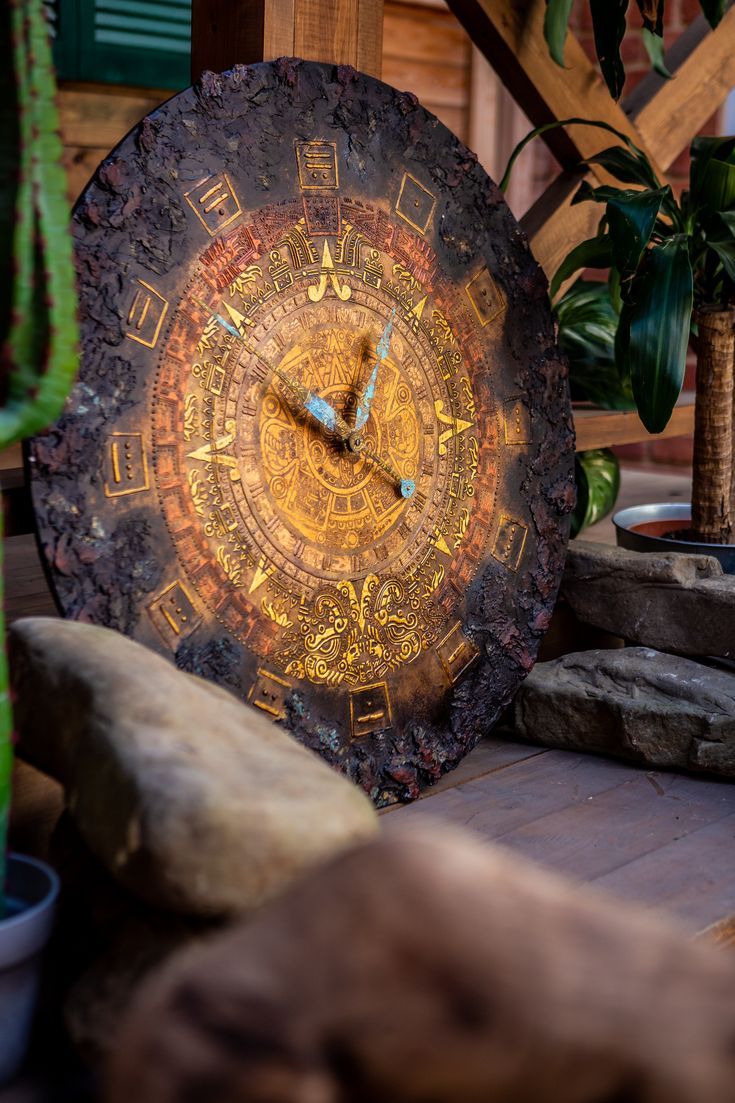 a large clock sitting on top of a wooden table next to rocks and plants in the background