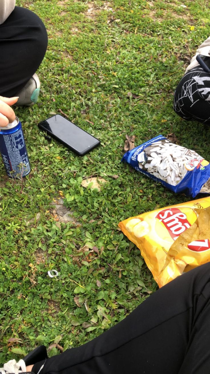 two people sitting on the grass with snacks and cell phones in front of their feet