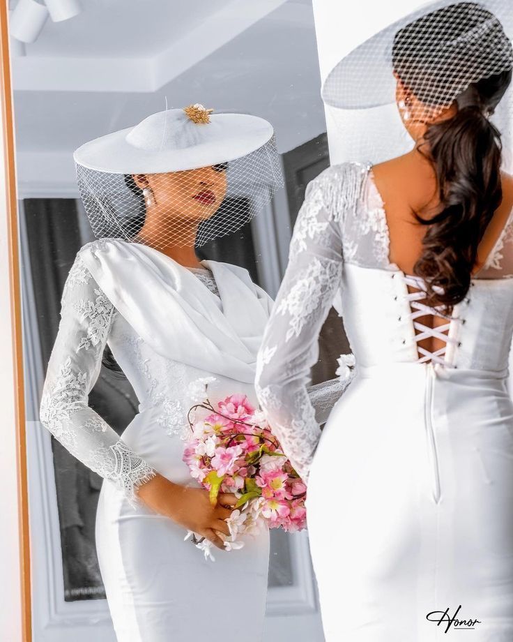 two women in white dresses and veils are looking at each other while one is holding flowers