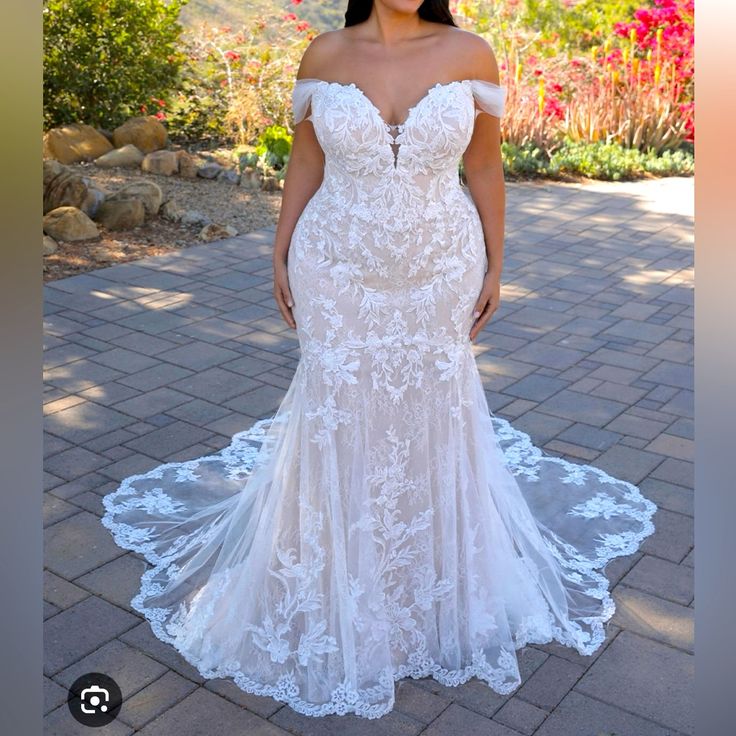 a woman in a white wedding dress standing on a brick walkway with flowers behind her