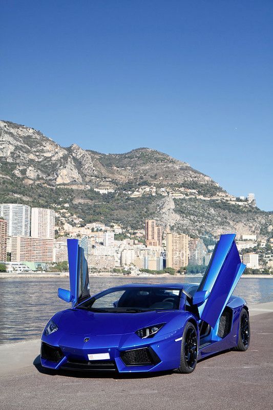 a blue sports car with open doors parked in front of a body of water and mountains