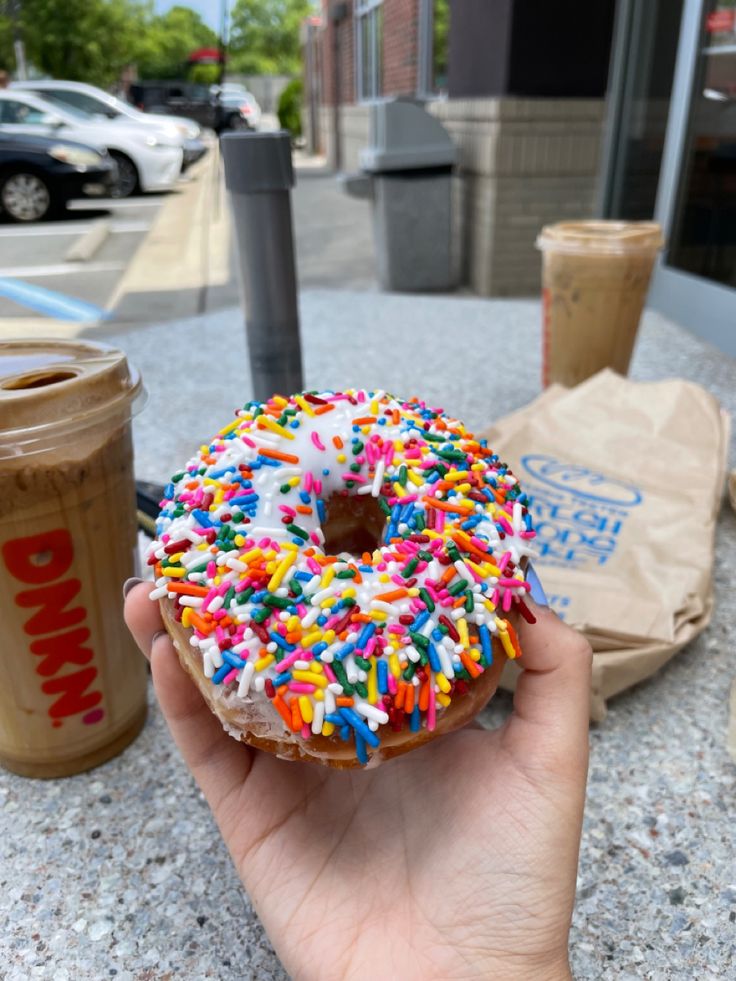 a hand holding a donut with sprinkles next to a cup of coffee