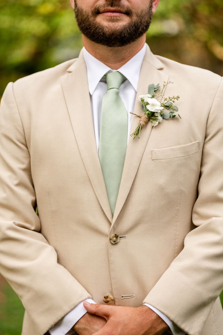 Close up picture of male wearing a tan suit jacket, with white shirt and sage green tie. Small green and white flowers wrapped in twine accent the right lapel Sage Groom, Rustic Wedding Groom Attire, Tan Suit Wedding, Tan Groomsmen, Groom Outfit Inspiration, Rustic Wedding Groom, Easton Maryland, Suit Inspiration, Green Wedding Suit