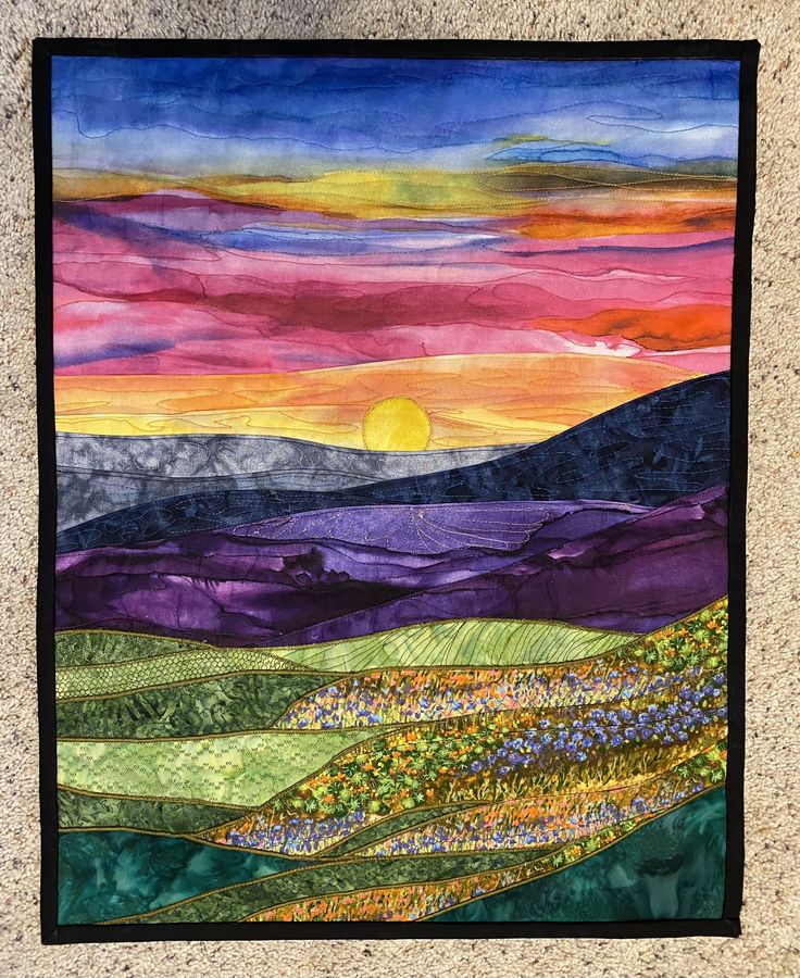 a quilted wall hanging on the side of a stone wall with a sunset in the background