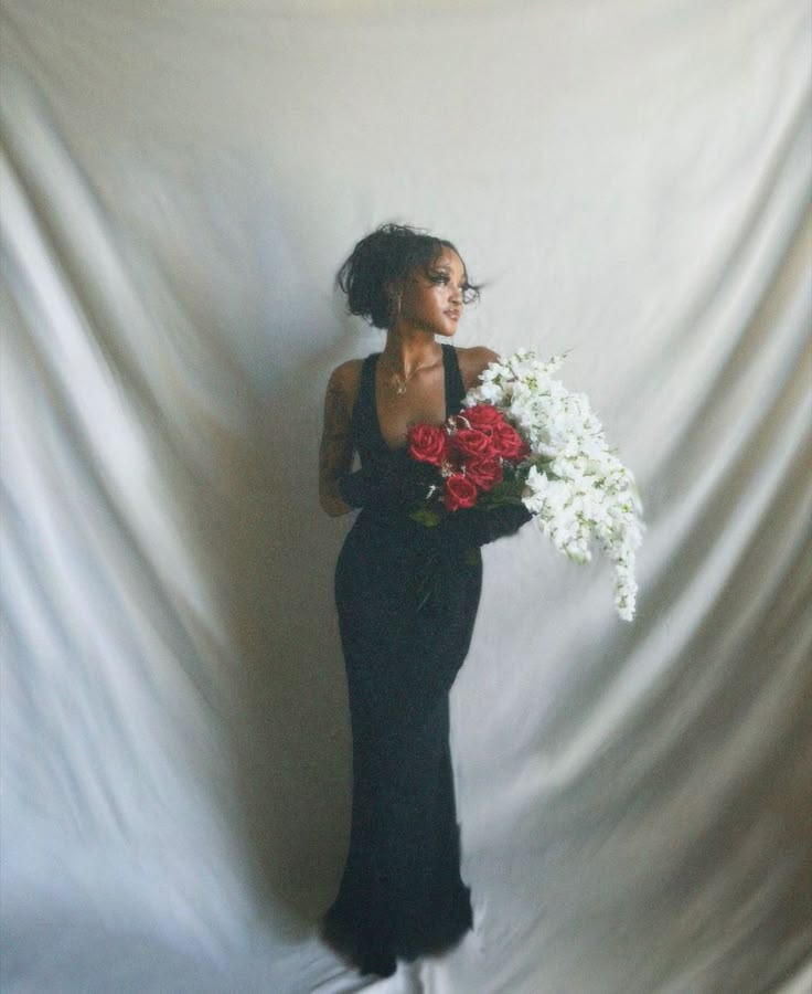 a woman in a long black dress holding white and red flowers with her hands behind her back