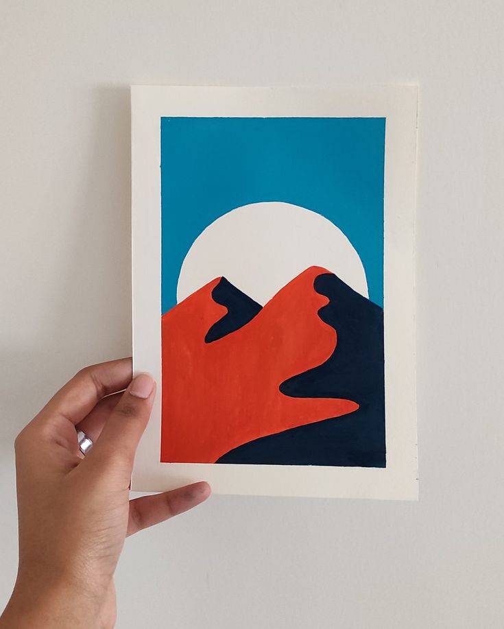 a hand holding up an art print with mountains and the moon in the sky behind it