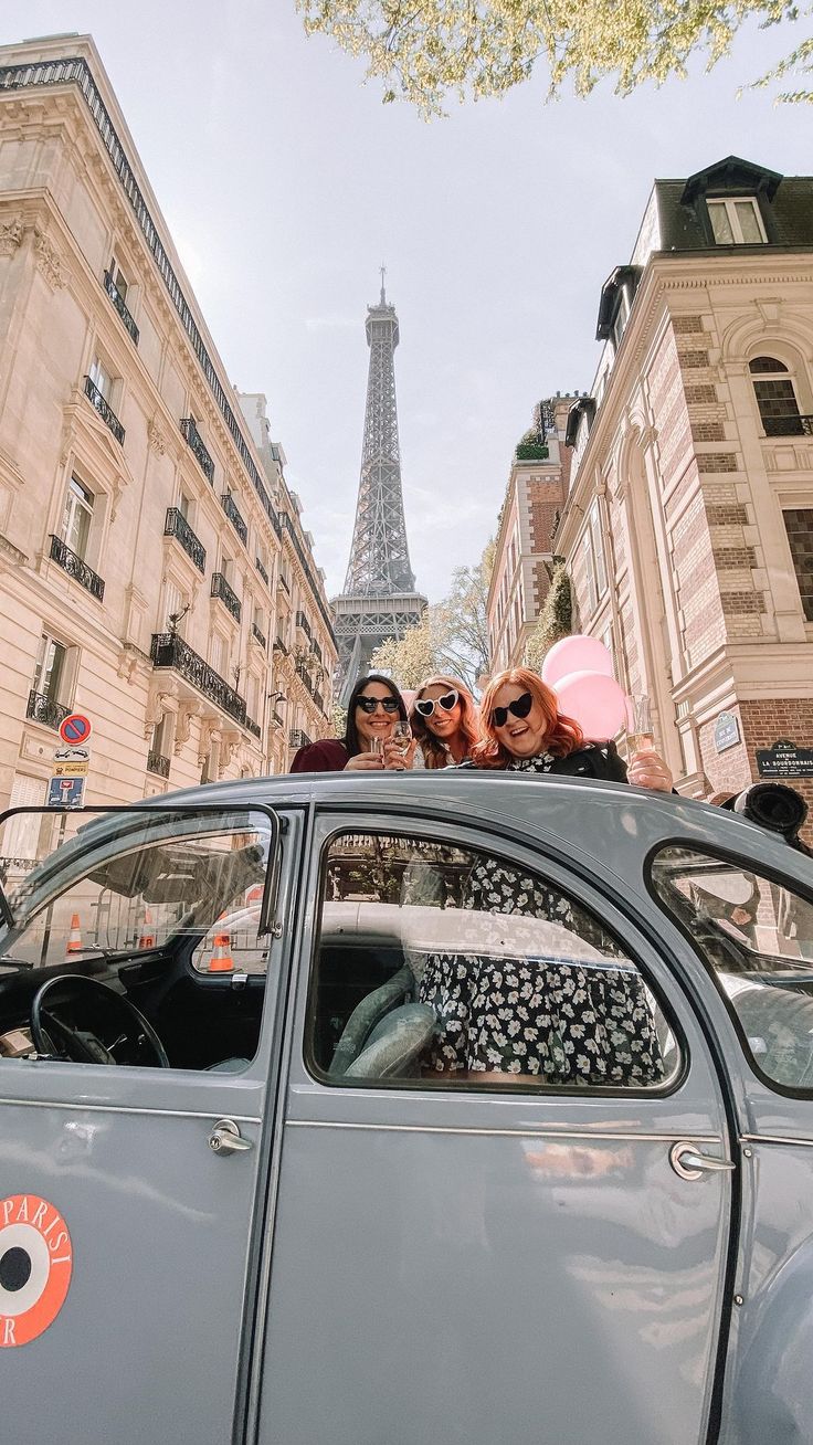 three women sitting on top of a car in front of the eiffel tower