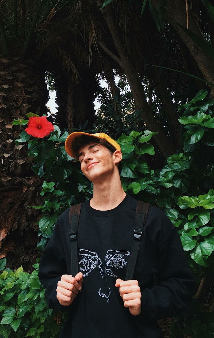 a man standing in front of some trees and plants with his eyes closed, wearing a hat