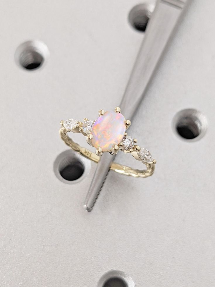 a white opal and diamond ring sitting on top of a piece of metal with holes in it