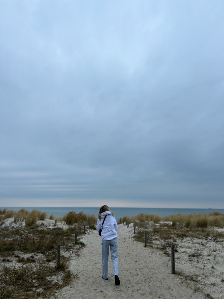 a woman walking down a sandy path towards the ocean on a cloudy day with a kite flying in the sky