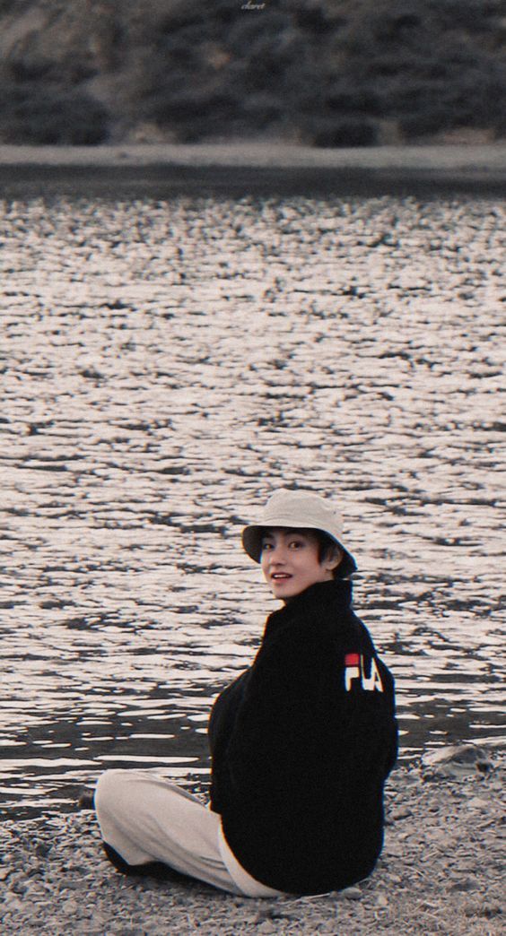 a young boy sitting on the ground next to a body of water wearing a hat