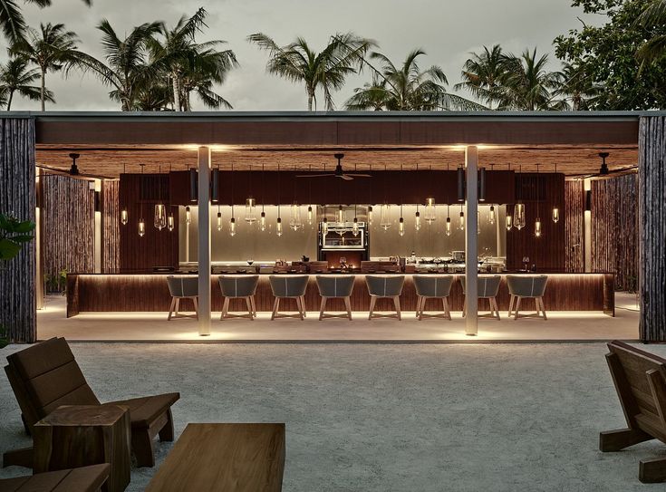 an outdoor bar with wooden chairs and lights on the outside, surrounded by palm trees