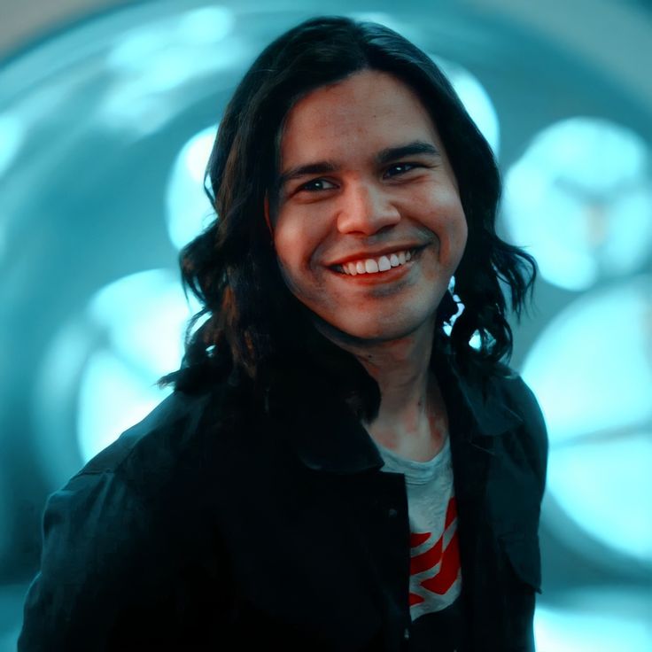 a man with long hair smiles at the camera while standing in front of a blue background