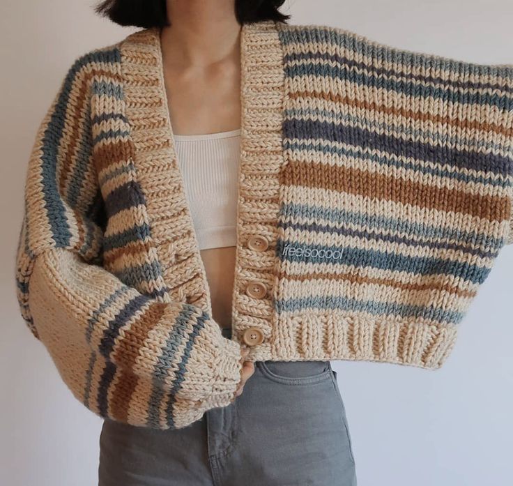 a woman wearing a striped sweater and cardigan with her hands in her pockets, standing against a white wall