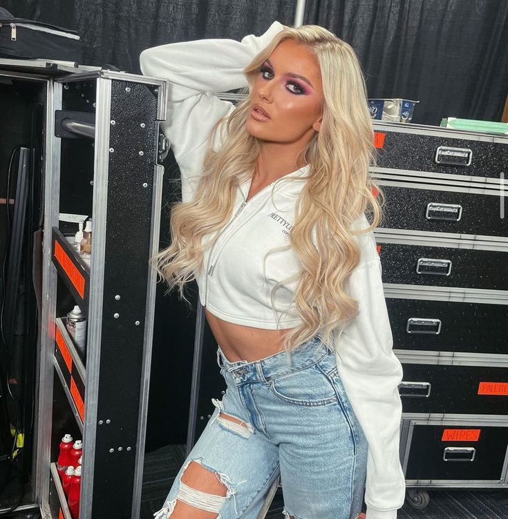 a woman with long blonde hair wearing ripped jeans and a white shirt standing in front of a tool cabinet