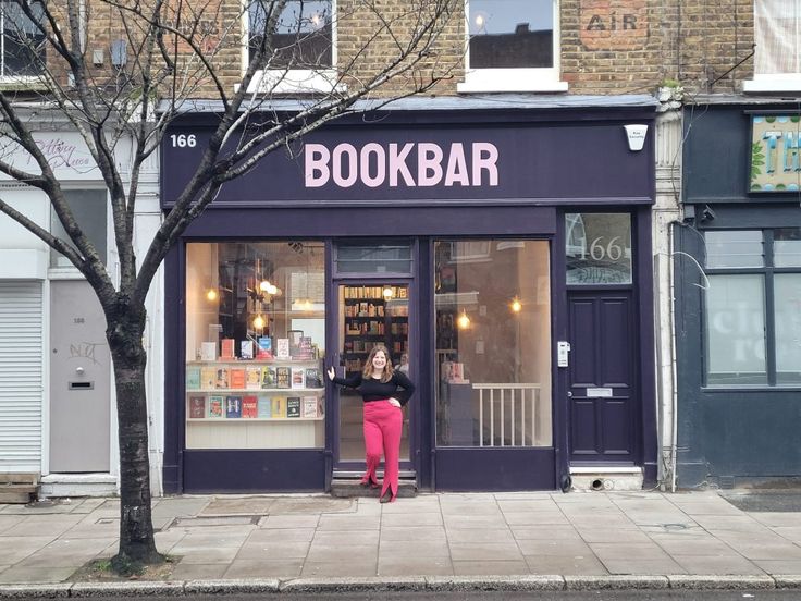 a woman standing in front of a book shop on the side of a street next to a tree