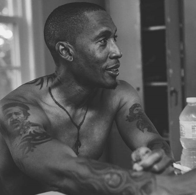 a man sitting at a table with tattoos on his arms and chest, holding a bottle of water in front of him