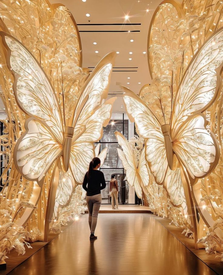 a woman walking down a hallway between two large gold butterfly sculptures in the middle of a building