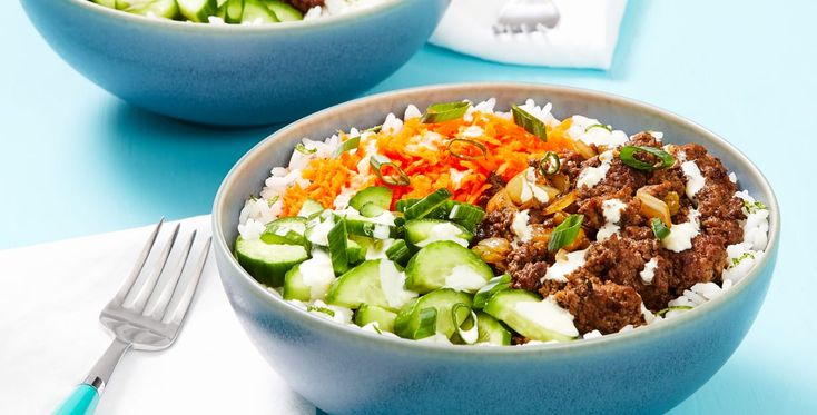 two bowls filled with meat and vegetables on top of a blue table next to silverware