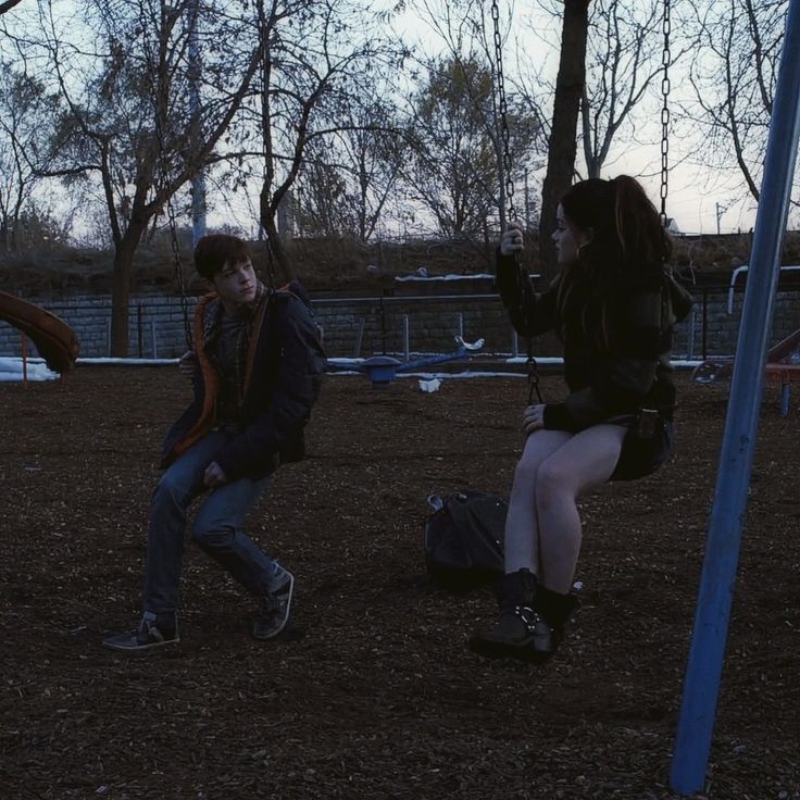 two people sitting on swings in a park at dusk, one is holding onto the other's back