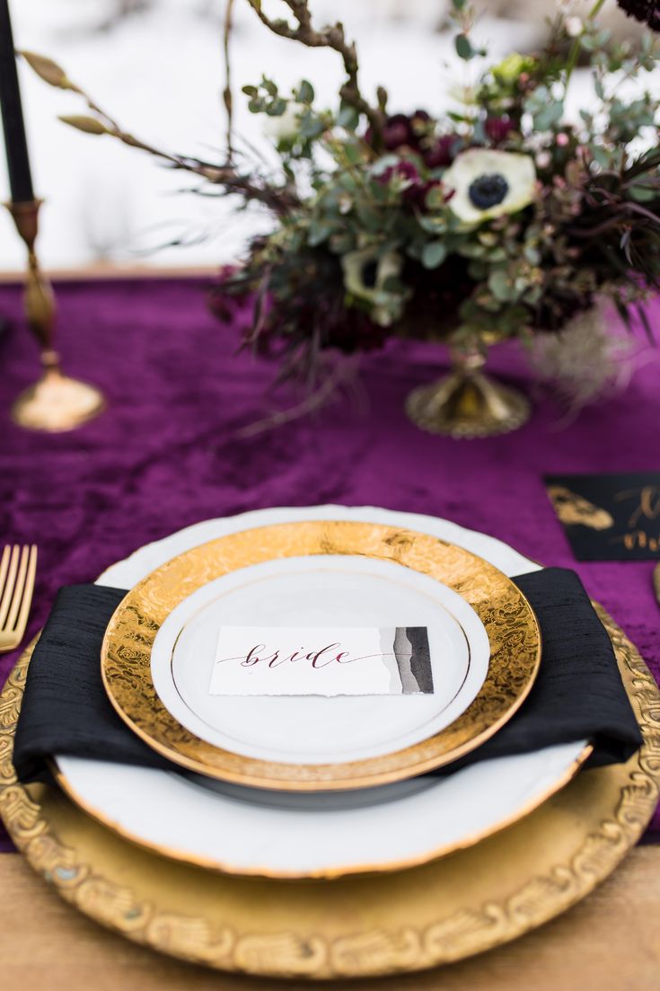 the table is set with black and gold plates, silverware, and purple napkins