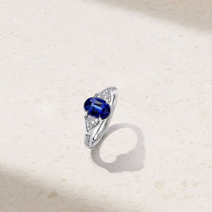 A modern statement piece, this ring showcases a three stone arrangement highlighted by a vibrant sapphire nestled amongst a pair of dazzling fancy cut diamonds. The center stone rests in prongs and is traced by a bezel-like border that extends and cradles the accompanying diamonds. Additional petite round diamonds cascade down along each side of the shank for additional sparkle. A true one-of-a-kind heirloom, wear yours solo for an impactful look. Metal: 18kt Gold Sapphire Weight: 2.18 ct. Fancy Stone Arrangement, Sapphire And Diamond Ring, Alexandrite Ring, Ruby Sapphire, Engagement Ring Wedding Band, High Jewelry, Three Stone, Wedding Ring Bands, Band Rings