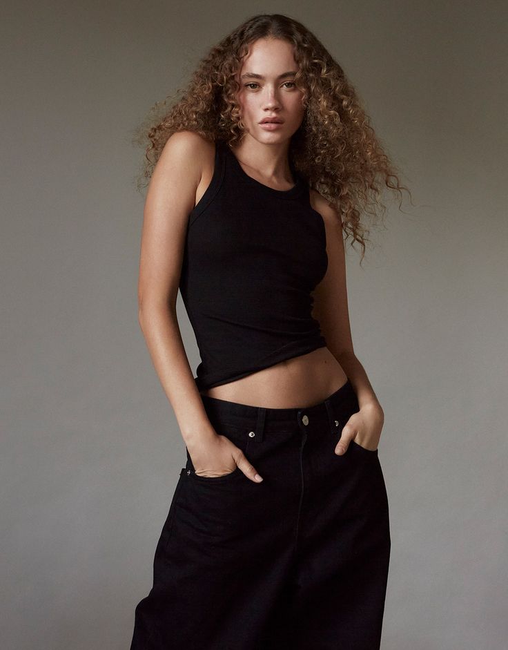 a woman with curly hair standing in front of a white wall wearing black pants and a crop top