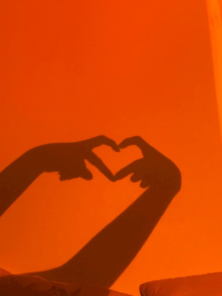 the shadow of two hands making a heart shape in front of an orange wall with pillows