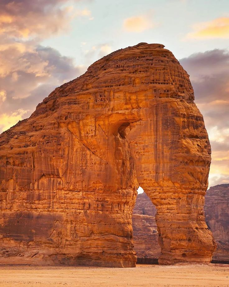 a large rock formation in the desert with a sky filled with clouds and sun shining through it