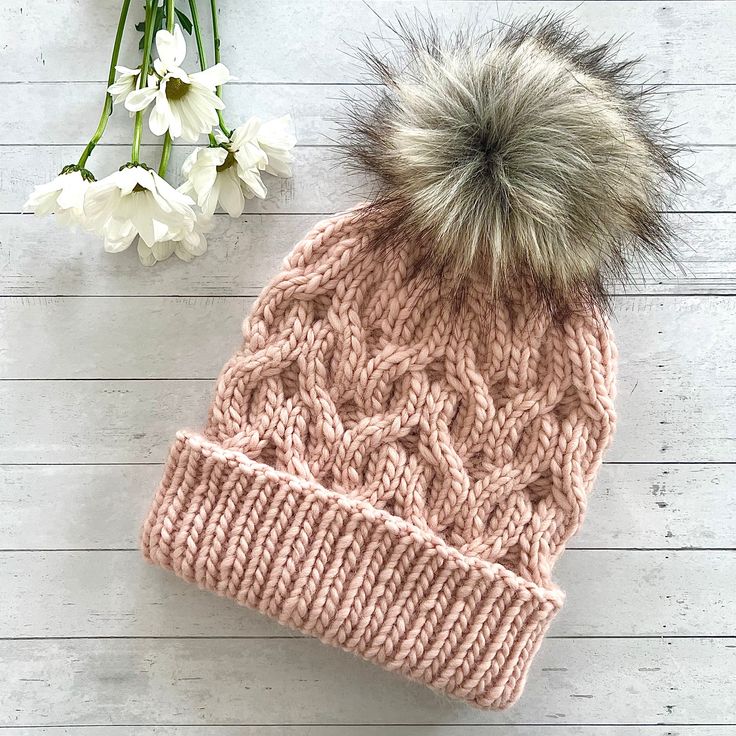 a knitted hat with a pom - pom sits next to some flowers