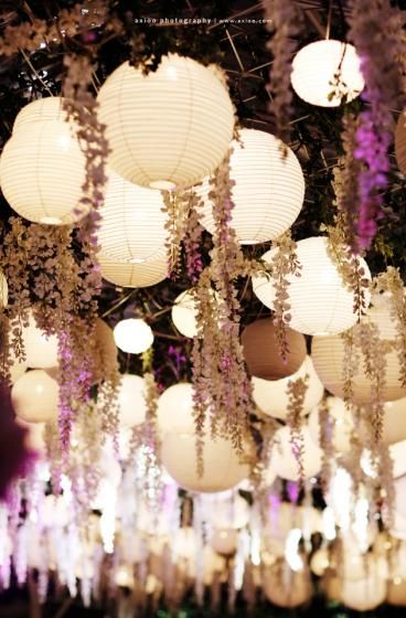 many white lanterns hanging from the ceiling with purple flowers and greenery all around them