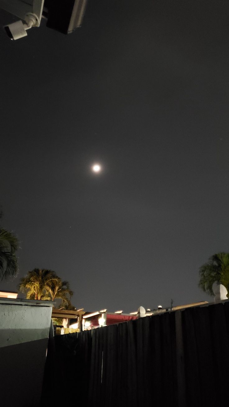 the moon is seen in the dark sky above some buildings and palm trees at night