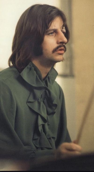 a man with long hair and a moustache looks off into the distance as he sits in front of a piano