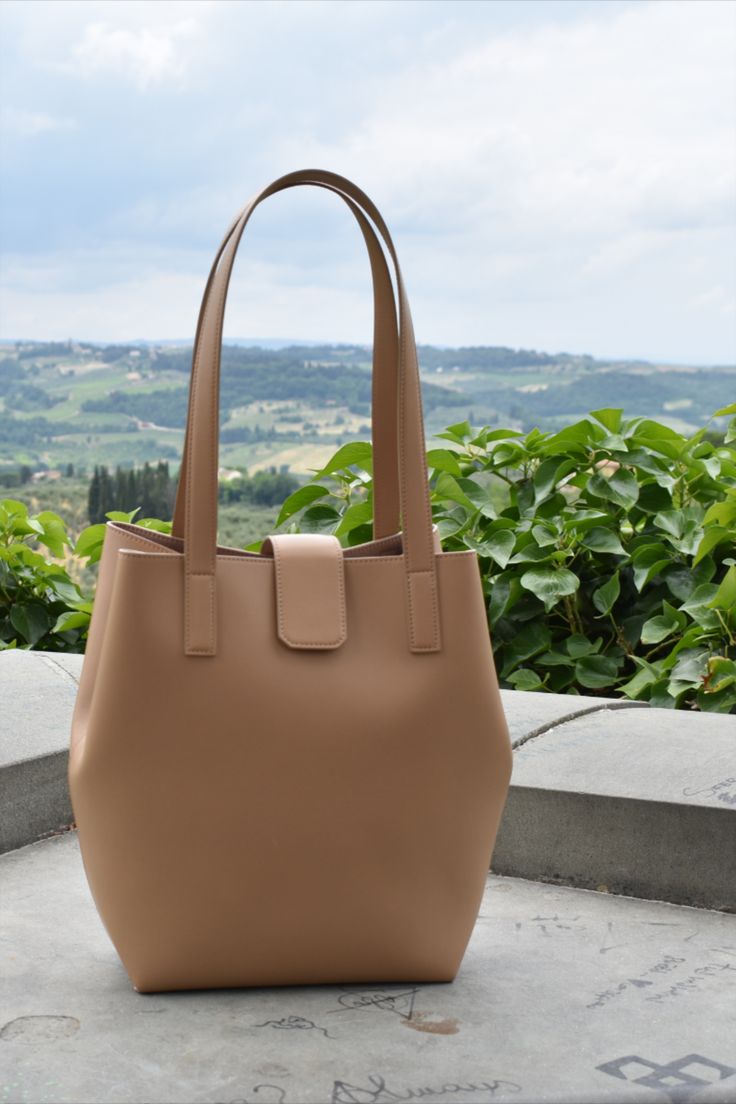 tan leather tote bag with tuscan hills in the background Chic Leather Bucket Bag With Magnetic Closure, Chic Bucket Bag With Smooth Grain, Chic Rectangular Bucket Bag With Smooth Grain, Versatile Everyday Bucket Bag With Magnetic Closure, Leather Bucket Bag With Smooth Grain For Evening, Everyday Bucket Bag With Magnetic Closure, Chic Smooth Grain Bucket Bag, Chic Top Handle Bucket Bag, Daily Use Bucket Bag With Magnetic Closure