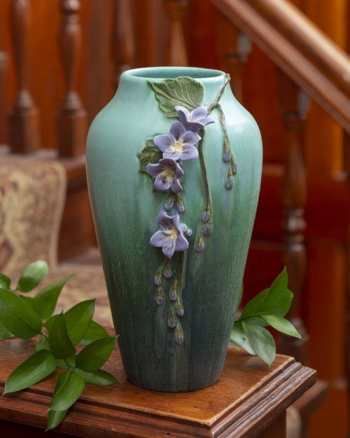 a green vase with purple flowers on it sitting on a table next to some stairs