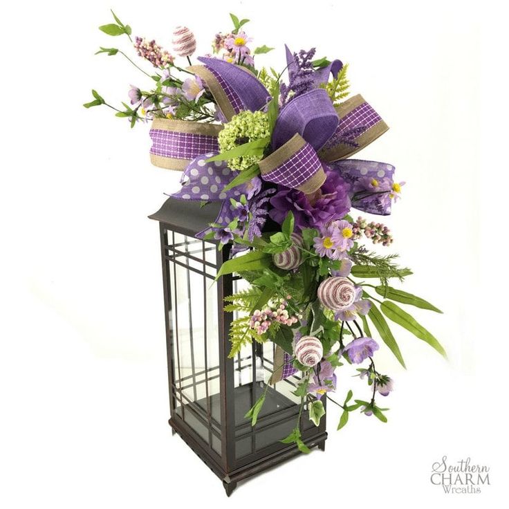 a purple and green arrangement in a black lantern with ribbons on it's sides