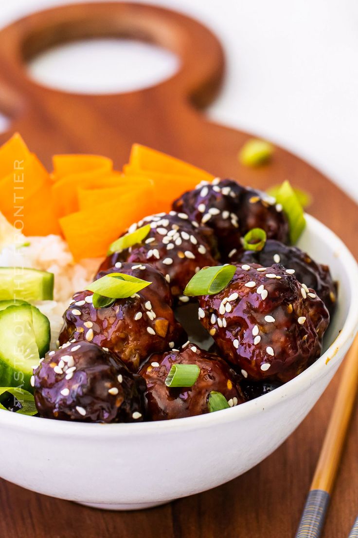 a white bowl filled with meatballs and vegetables next to chopsticks on a wooden cutting board