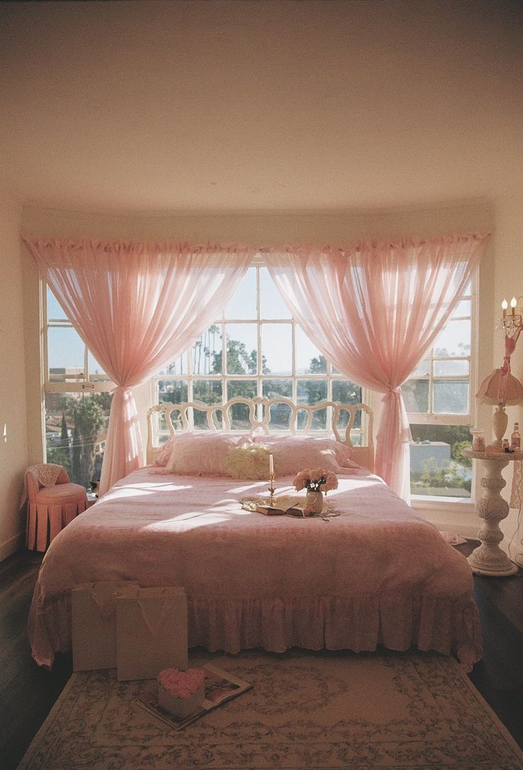 a bed with pink sheets and pillows in front of a large window that has sheer curtains on it